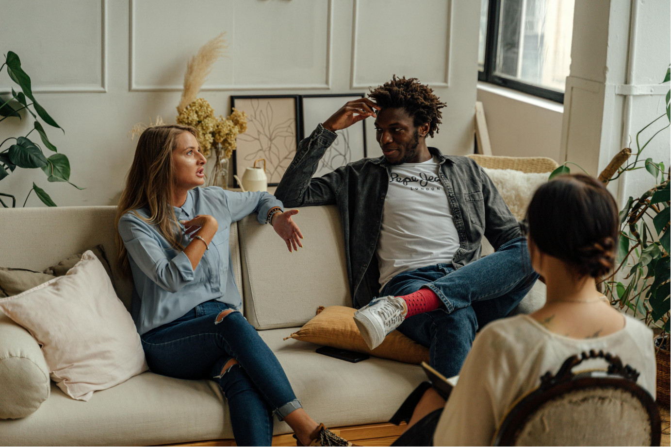 A couple in an emotional conversation on a couch while their therapist listens and takes notes nearby