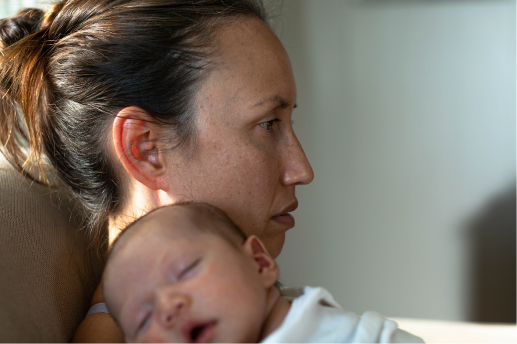 A mother's profile, looking pensive while holding her sleeping newborn against her shoulder.